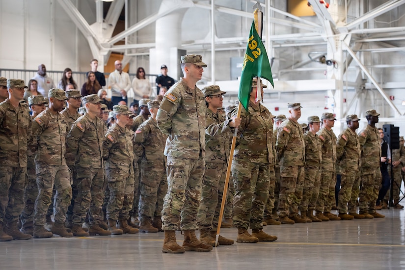 Men and women dressed in camouflage military uniforms stand in formation. A man at the front of the formation holds a wooden pole with a green flag attached to the top of it while two people in similar attire stand next to him.