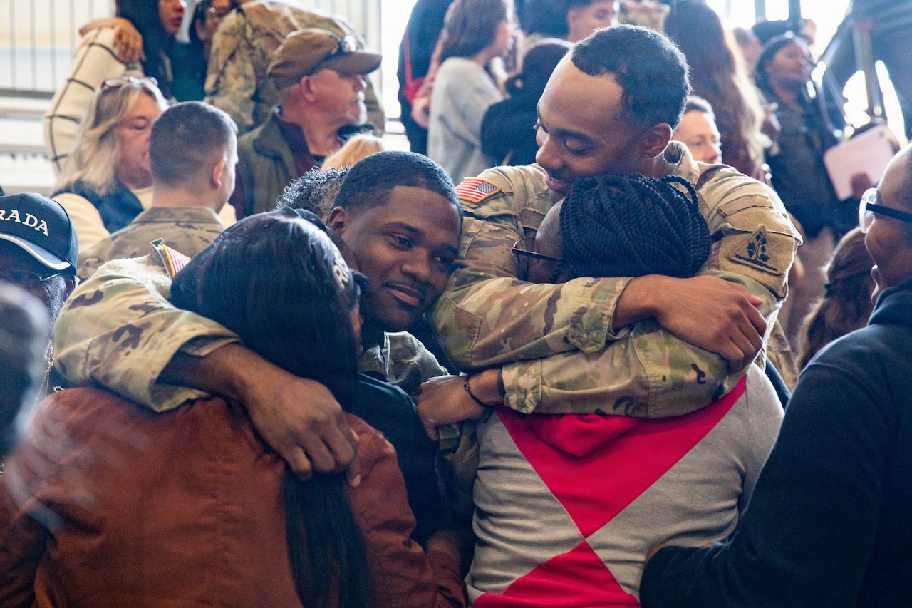 Two men in camouflage military uniform hug two women as others look on.