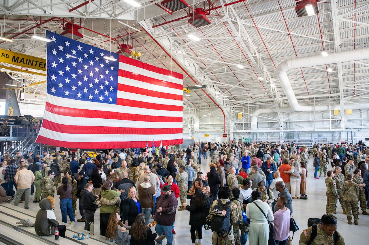 A crowd of people congregate in an aircraft hangar; some of them are wearing camouflaged military uniforms. A large American flag is hanging from the roof.