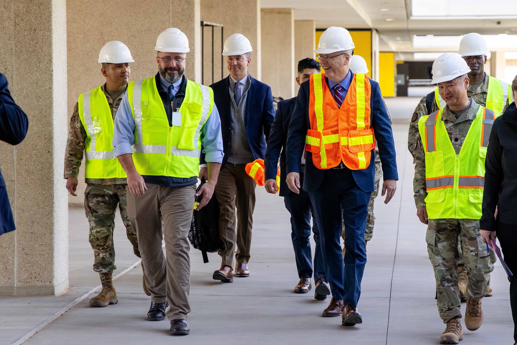 A group of men in Air Force uniforms and suits walk on the sidewalk next to a building while wearing safety vests and hard hats.