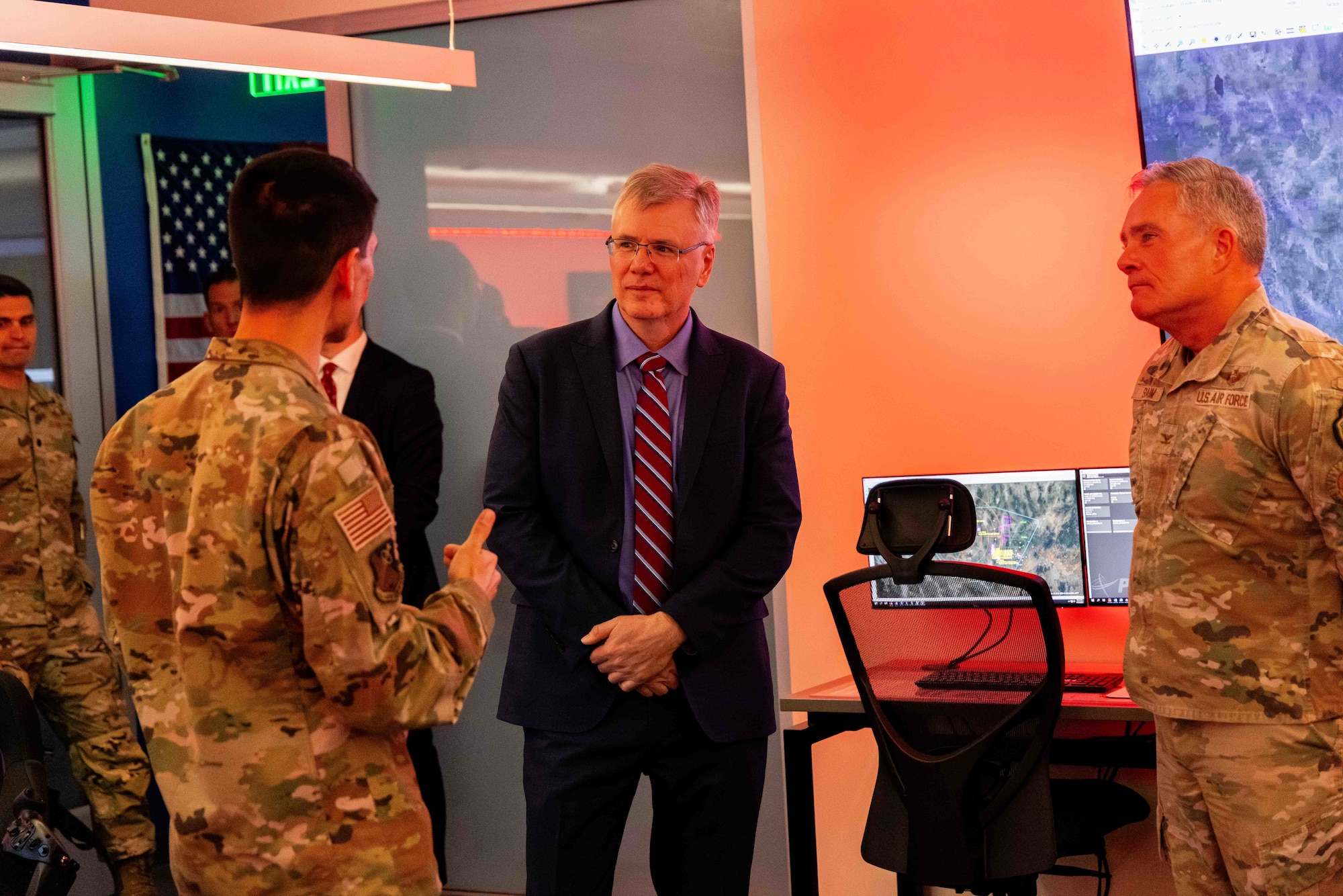 Two Airmen in uniform talk with the Secretary of the Air Force inside of a red tinted room.