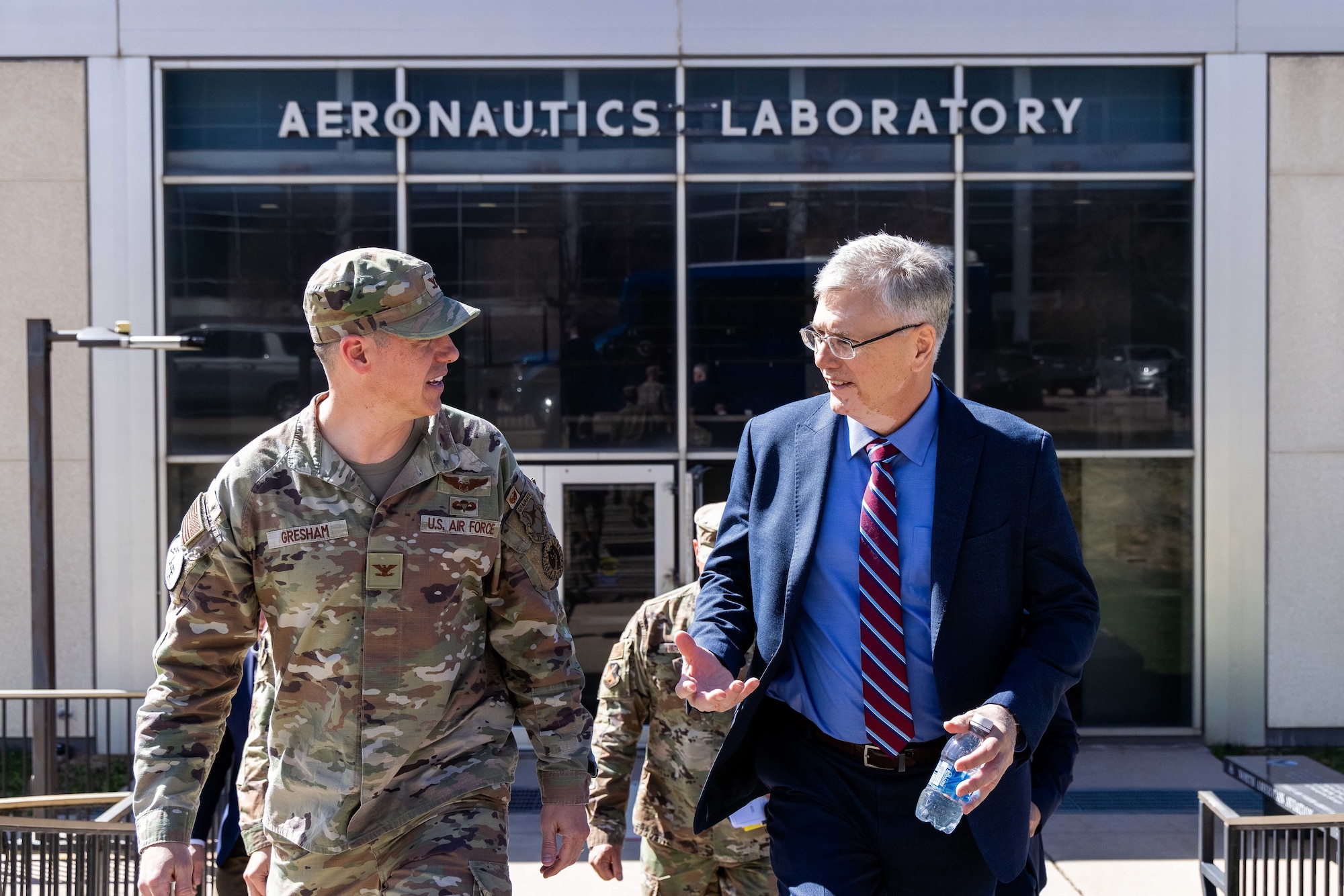 A man in a suit and an Airman in his camo uniform walk away from the entrance of an aeronautics laboratory building.