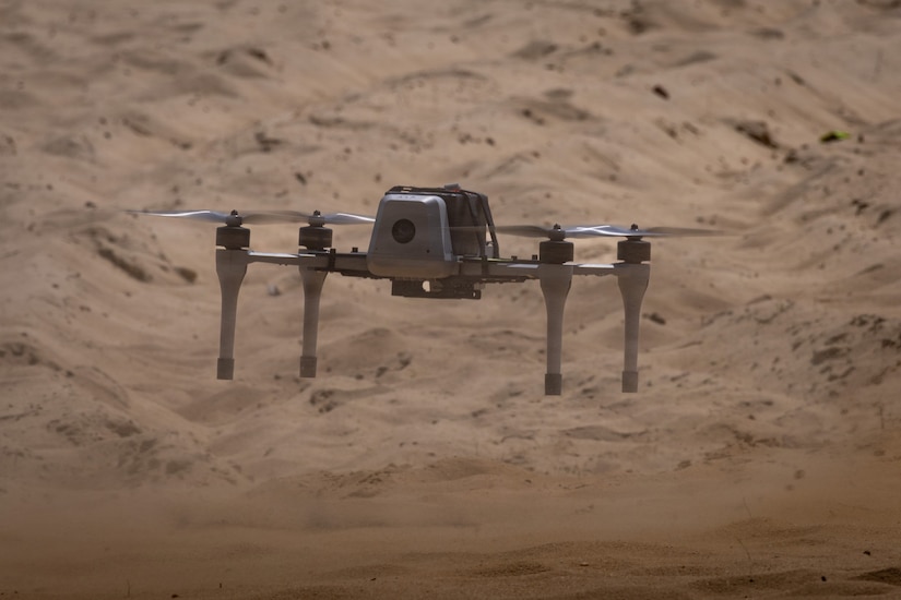 A small, unmanned drone with four propellers and a camera sits on the ground in a desert terrain before it takes off.