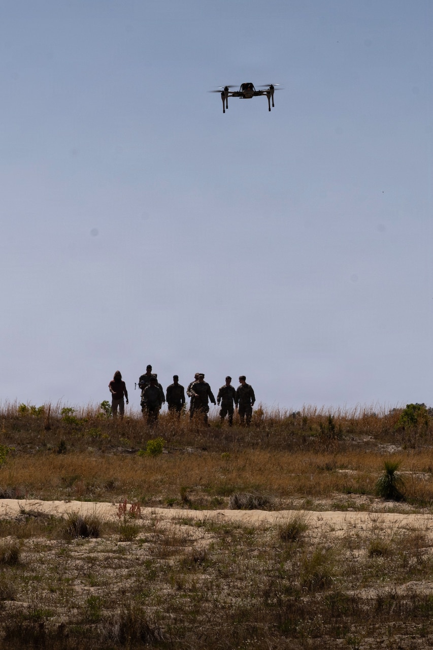 The silhouettes of several people standing in tall grass watch as a drone flies in a blue sky.