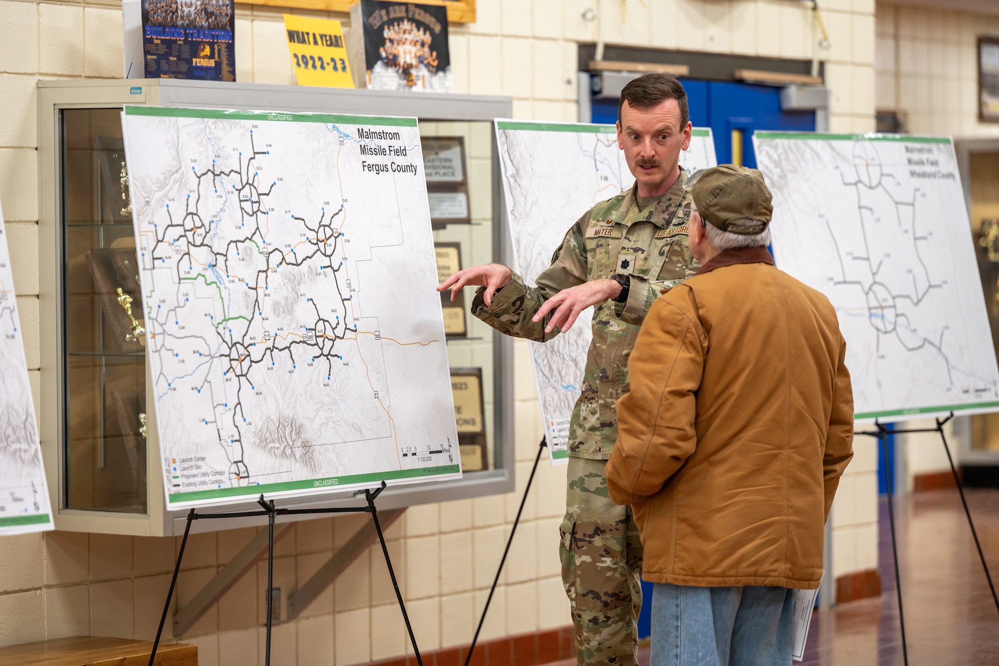 A man in uniform talking with another man in front of displayed maps.