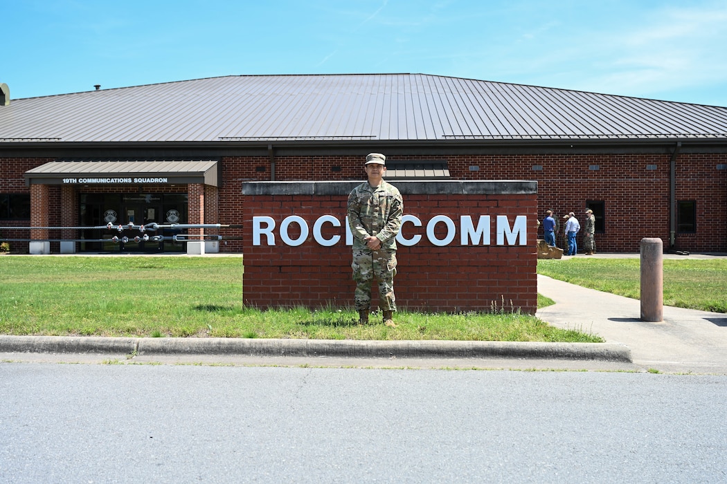 A man is posing for a picture in front of a "Rock Comm" sign.