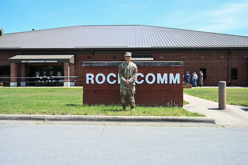 A man is posing for a picture in front of a "Rock Comm" sign.