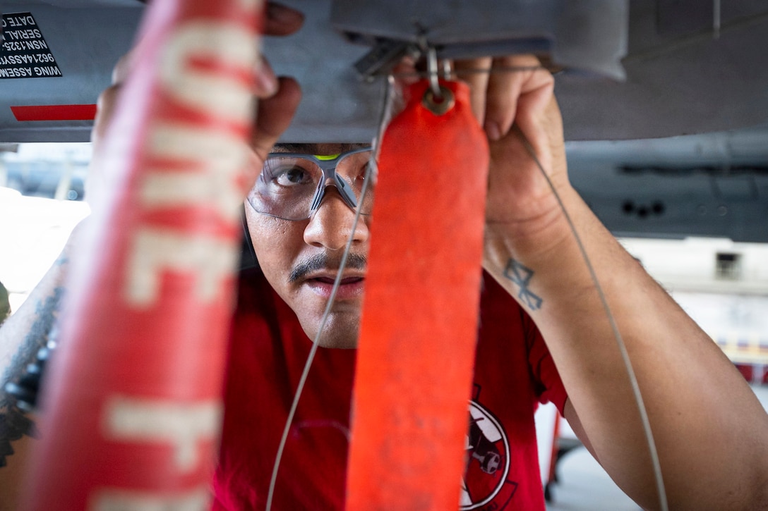 A close-up of an airman wearing a helmet and clear glasses adds wire to an aircraft during the day.