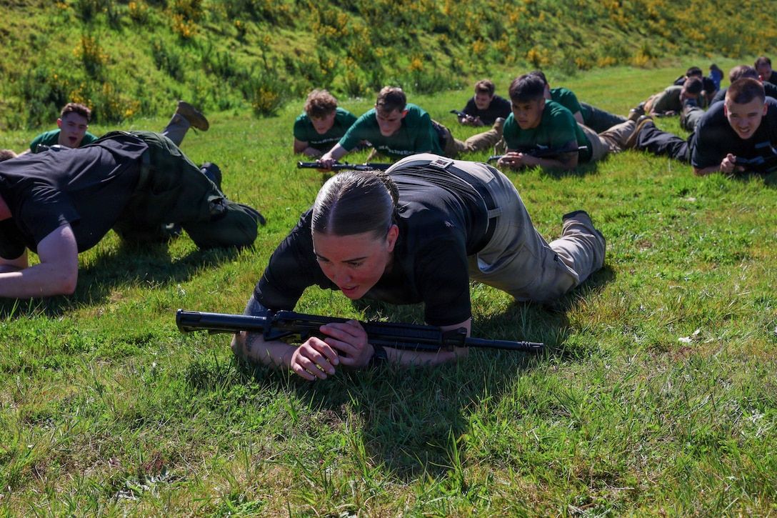 A Marine Corps officer candidate crawls on a field with a weapon as a group of kids follow behind.