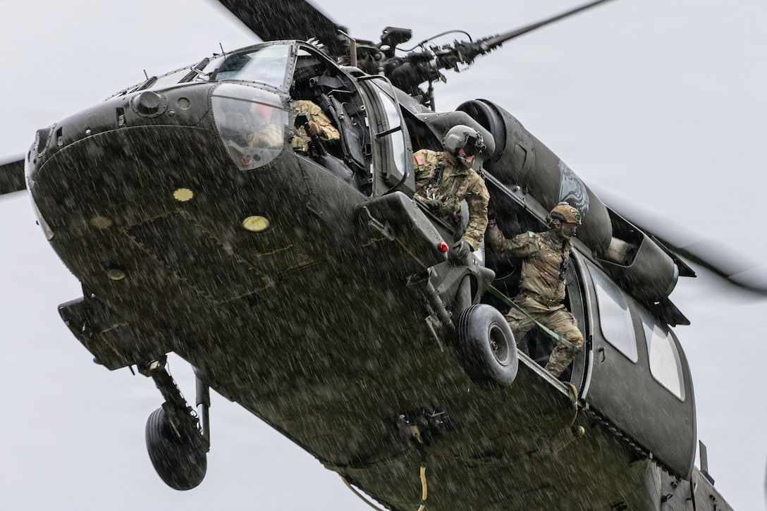Soldiers look out the window of a helicopter as it flies into a gloomy sky.