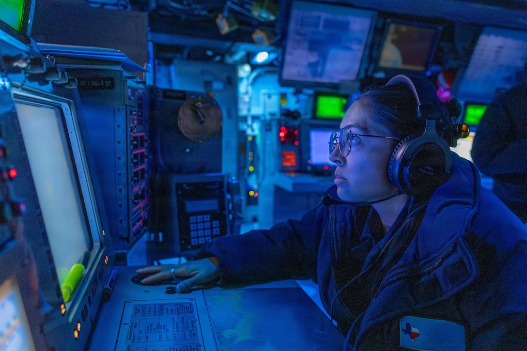 A sailor wearing headphones works in front of a screen in a dark control room illuminated by blue light.