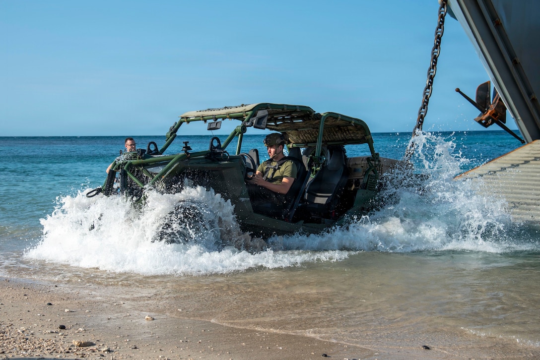 A Marine in a vehicle drives into the water on the shore of a beach from a vessel's ramp.