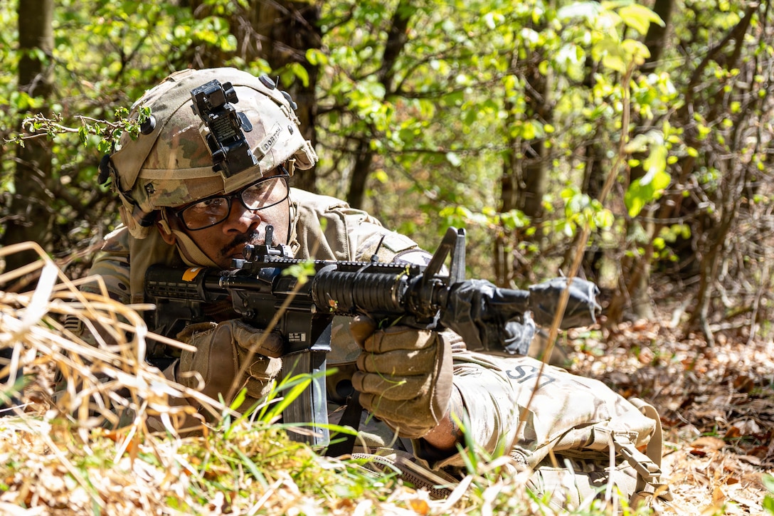A soldier points his machine gun while on the ground in a wooded area.