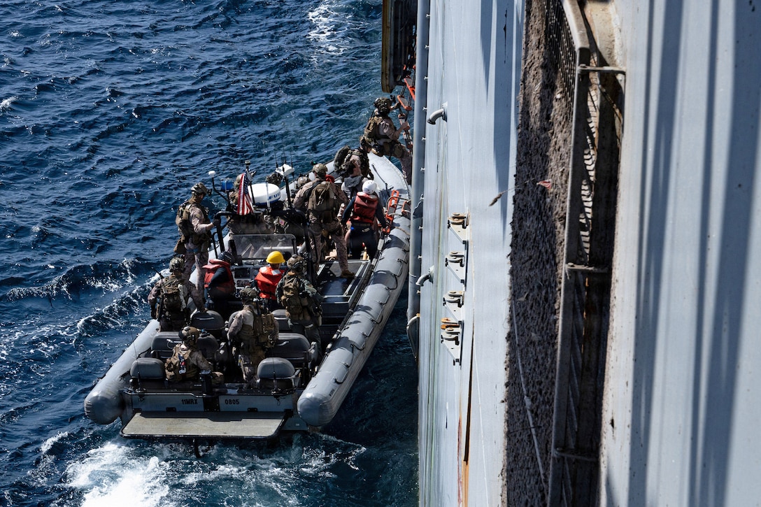 Marines on a small boat get in line and begin to climb onto a large vessel.
