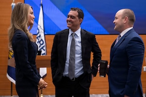 A woman and two men, all in business attire, stand and speak in a conference room with a screen and two flags in the background.