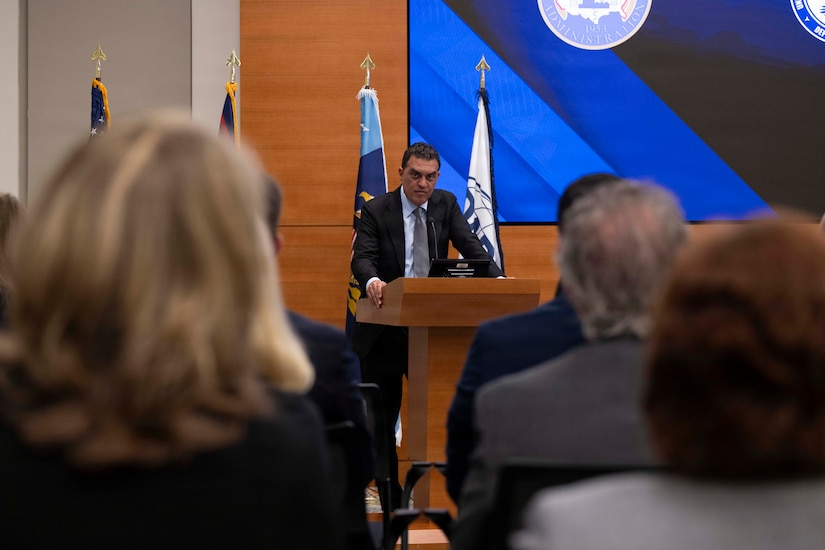 A man in business attire stands behind a lectern and speaks to an audience; behind him are various flags and a screen.