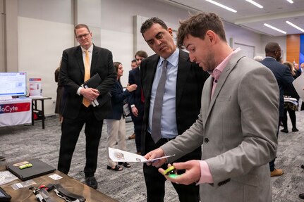 Two men in business attire stand near each other and look at a sheet of paper. In front of them is a table with various pieces of technology on it, and in the background are people in similar attire talking.