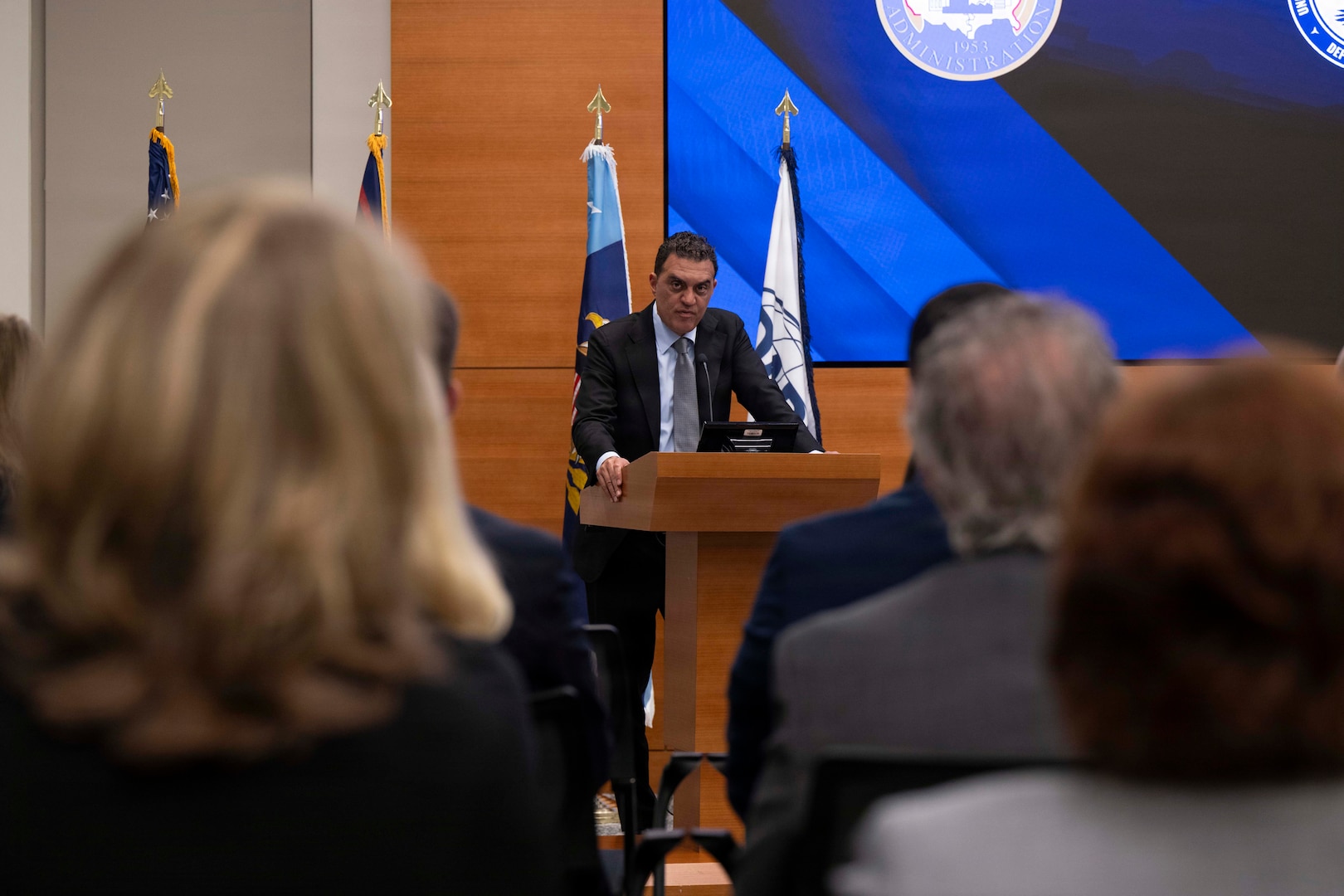 A man in business attire stands behind a lectern and speaks to an audience; behind him are various flags and a screen.