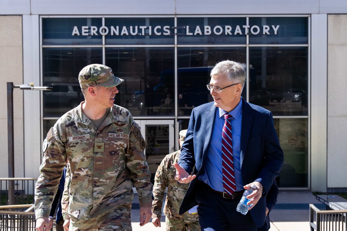 A man in a suit and an Airman in his camo uniform walk away from the entrance of an aeronautics laboratory building.