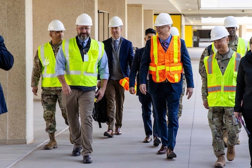 A group of men in Air Force uniforms and suits walk on the sidewalk next to a building while wearing safety vests and hard hats.