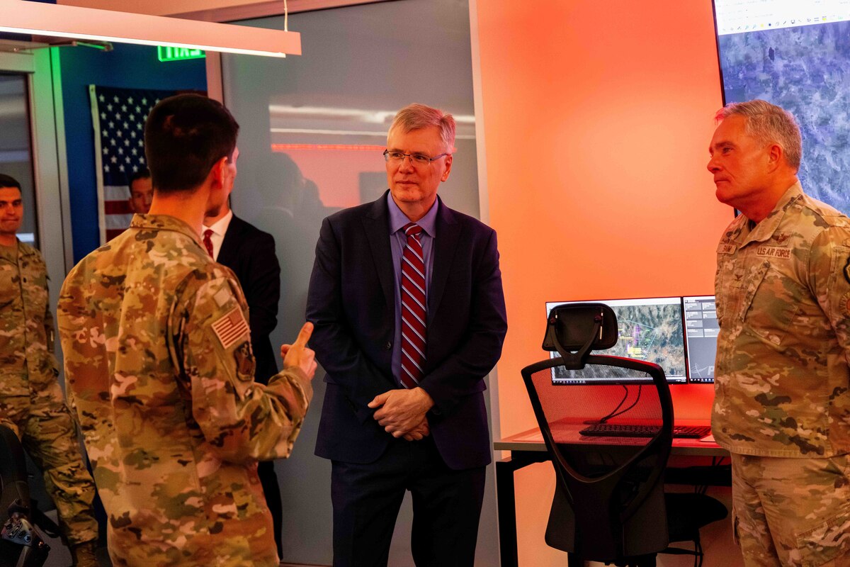 Two Airmen in uniform talk with the Secretary of the Air Force inside of a red tinted room.