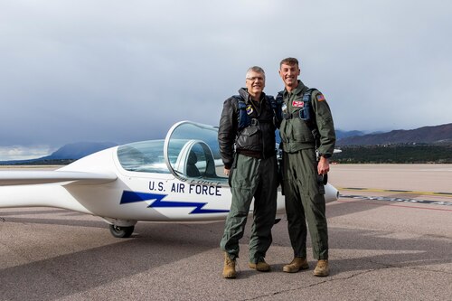 Two men in flight suits pose for a photo in front of a glider.