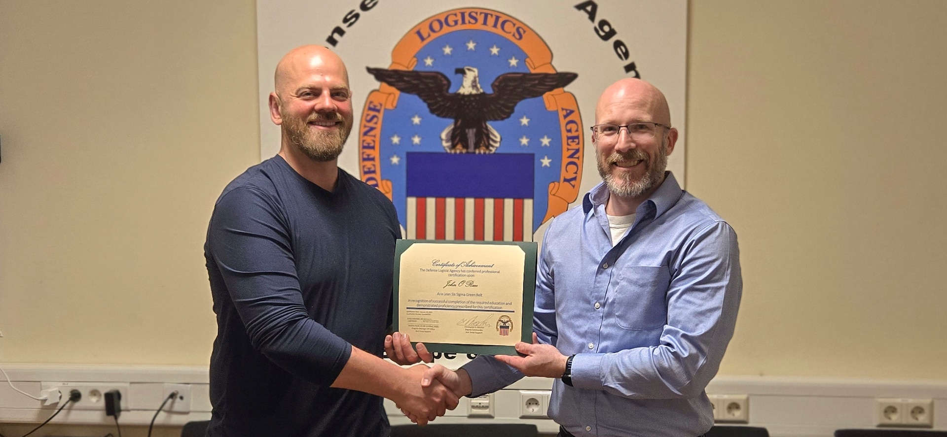 John Race, left, receives a Lean Six Sigma Green Belt Certificate of Achievement from his supervisor, Keith R. Smoly, Defense Logistics Agency information Operations regional director, in Sigonella, Italy, April 30, 2026. Race led a continuous process improvement project for DLA Distribution that successfully streamlined a recurring weekly task. The initiative utilized long-distance coaching from DLA Troop Support to create a standardized, efficient workflow that can be adopted across the enterprise.