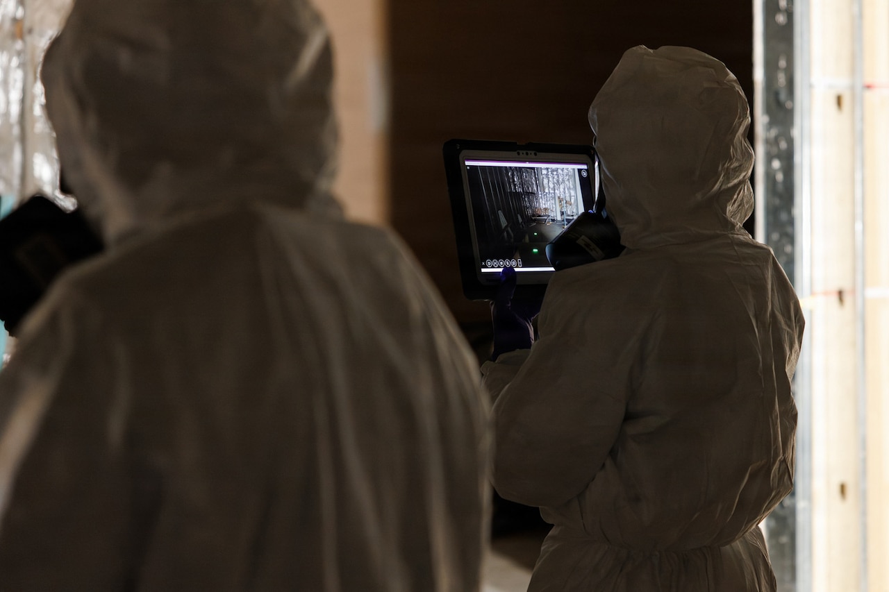 A person in a hazardous material suit takes pictures on a tablet while looking at an empty room; another person in similar attire stands next to them.