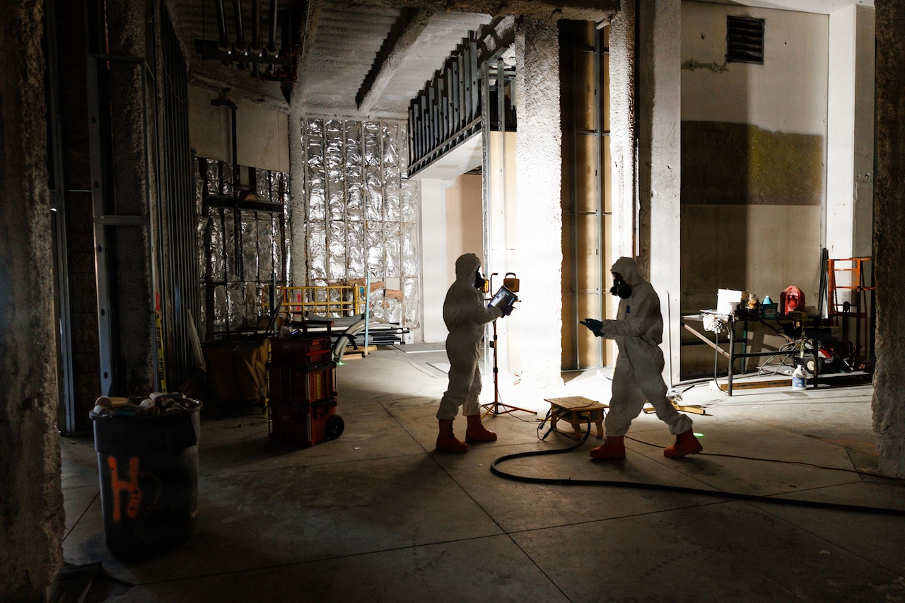 Two people in hazardous material suits monitor an empty room while holding tablets in their hands.