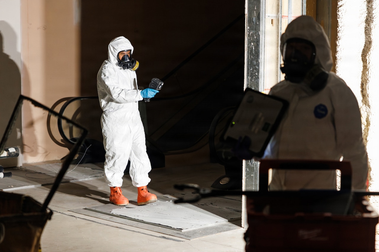 Two people in hazardous material suits monitor an empty room. One of the people holds a radiation detection device while the other person holds a tablet.
