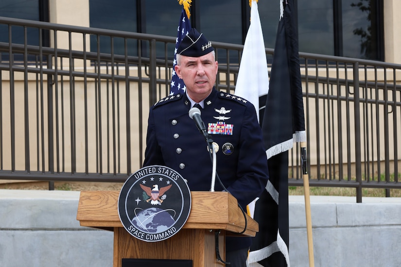 A man wearing a military dress uniform stands behind a lectern speaking into a microphone; there are three flags and a large building behind him. An emblem on the lectern reads, “United States Space Command.”