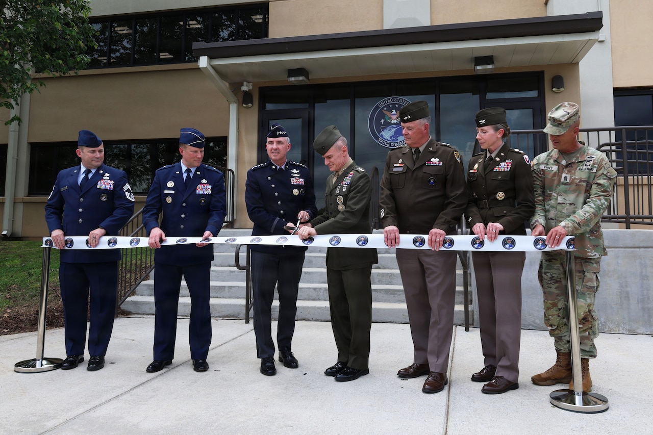 Two men wearing military dress uniforms use a pair of large scissors to cut a ribbon in front of a building while five people in similar attire hold the ribbon as they stand on the sides of the two men. An emblem of the building reads, "United States Space Command."