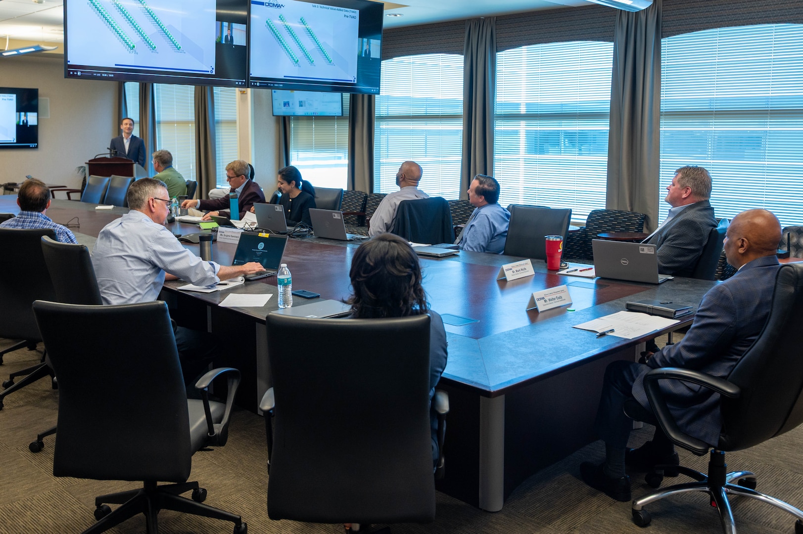 People sitting at a table watching presenter and presentation screens.