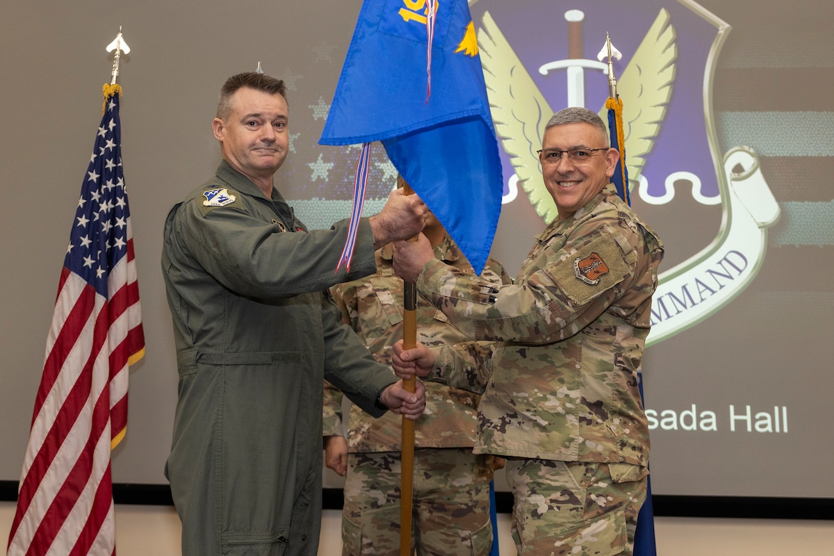 Two military members stand facing the camera, holding a guidon together. A military member stands at attention behind them.