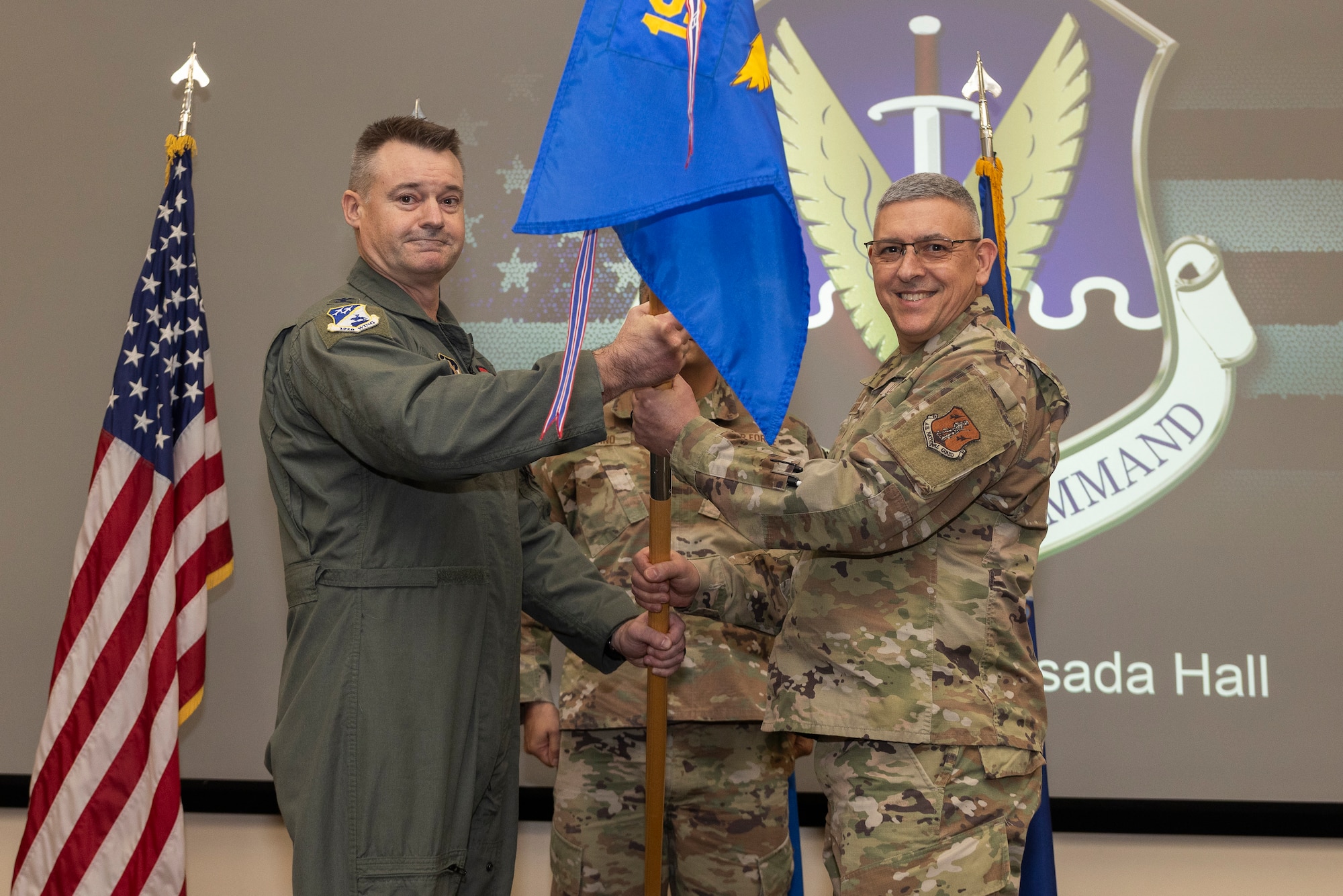 Two military members stand facing the camera, holding a guidon together. A military member stands at attention behind them.