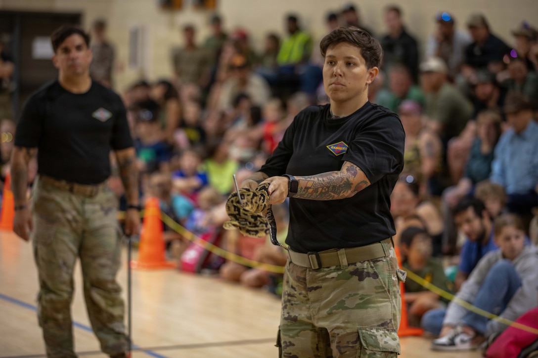 A soldier holds a snake as a fellow soldier watches in front of a blurred, seated audience.