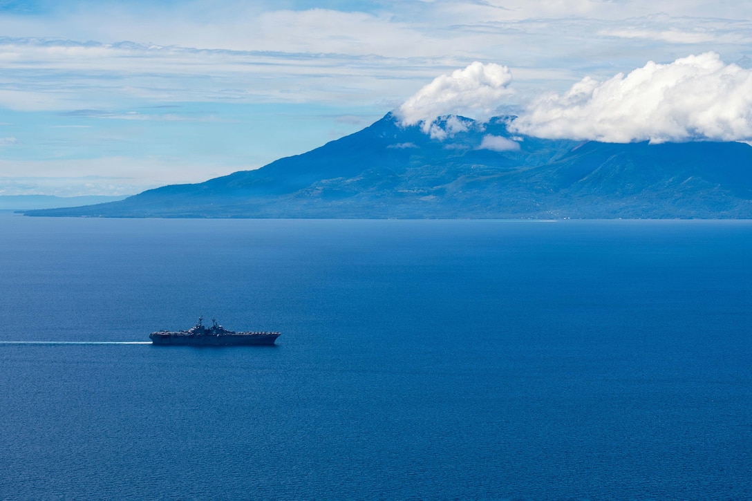 A ship transits a body of water under a blue sky with a mountain in the background.