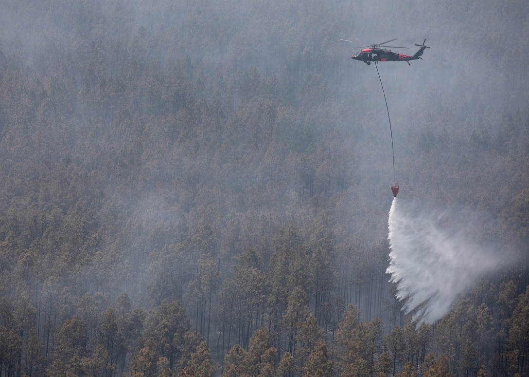 An airborne helicopter uses a bucket to spray water on a wooded area during the day.