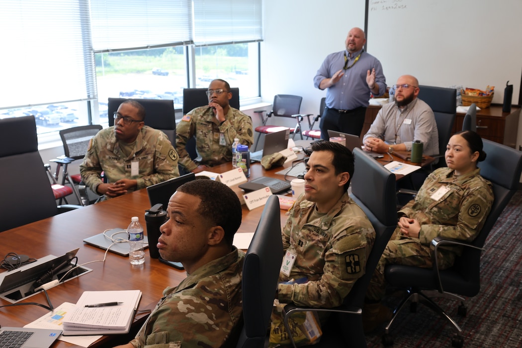 Several uniformed Soldiers and Sailors sitting at a conference table as Huntsville Center Medical program manager provide an information slide show on various programs.