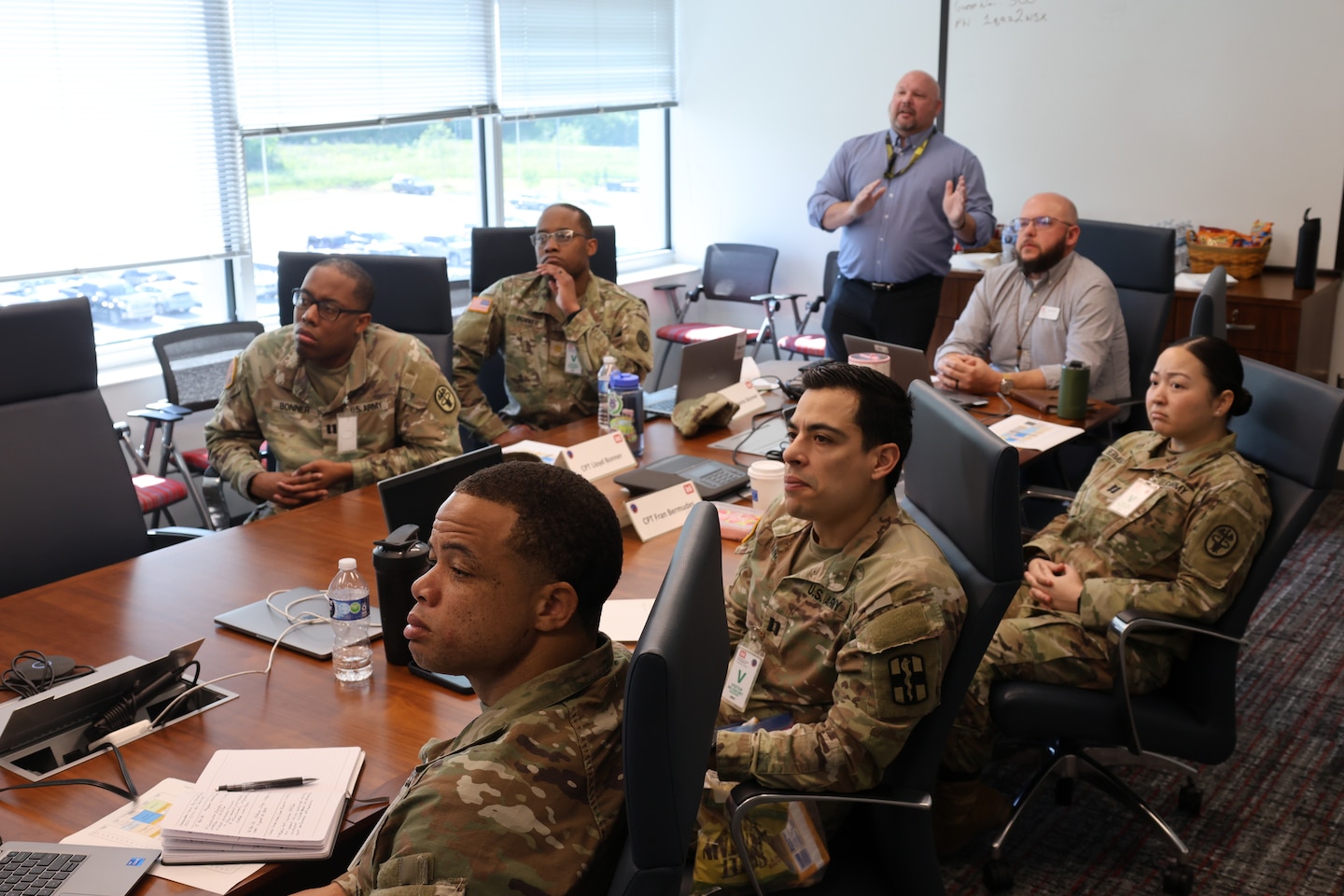 Several uniformed Soldiers and Sailors sitting at a conference table as Huntsville Center Medical program manager provide an information slide show on various programs.