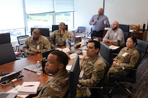Several uniformed Soldiers and Sailors sitting at a conference table as Huntsville Center Medical program manager provide an information slide show on various programs.
