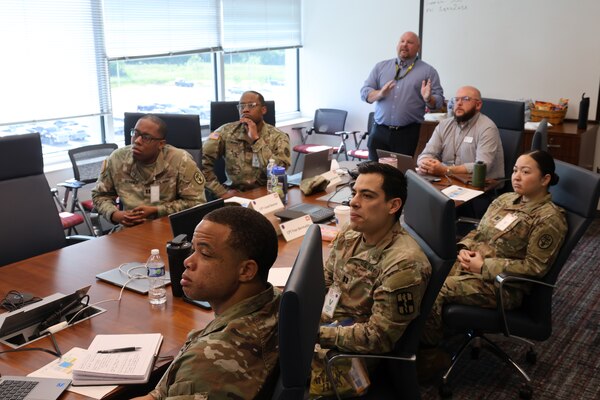 Several uniformed Soldiers and Sailors sitting at a conference table as Huntsville Center Medical program manager provide an information slide show on various programs.