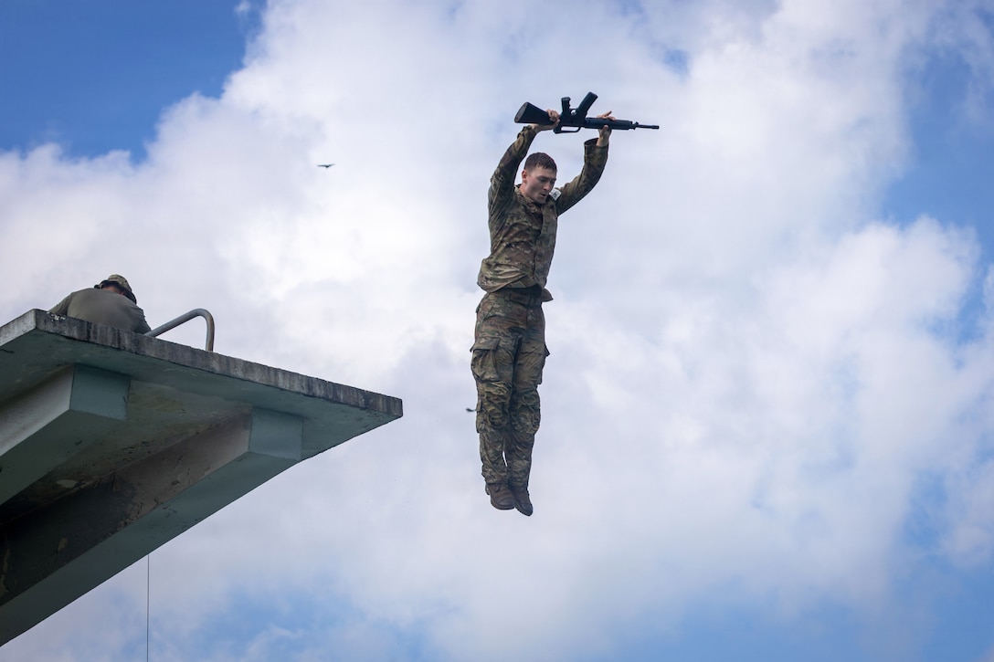 A soldier holds a weapon in the air while descending against a cloudy, blue sky with a diving board to the left.