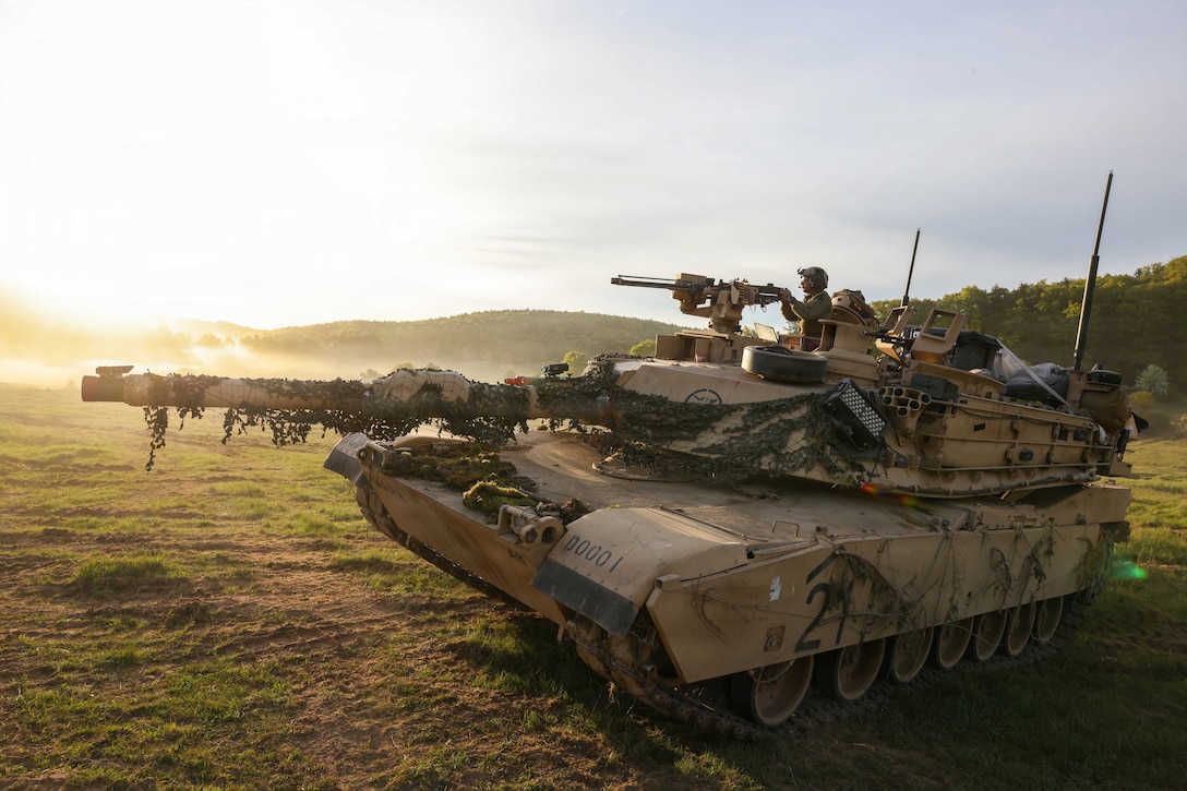 A soldier operates a tank on a field as the sun shines brightly from the top left corner.
