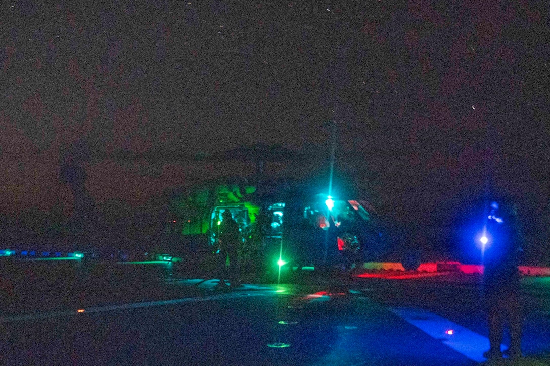 Marines walk on a dark flight deck near a helicopter illuminated with red, green and blue lights.