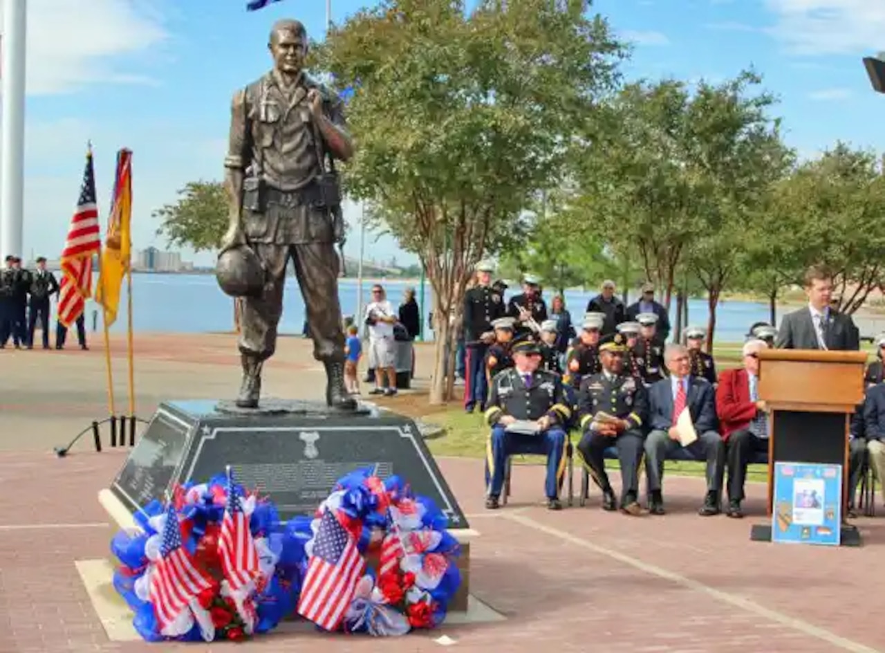 A statue of a soldier stands atop a pedestal as a man in a suit speaks to a gathering of military and civilian people.