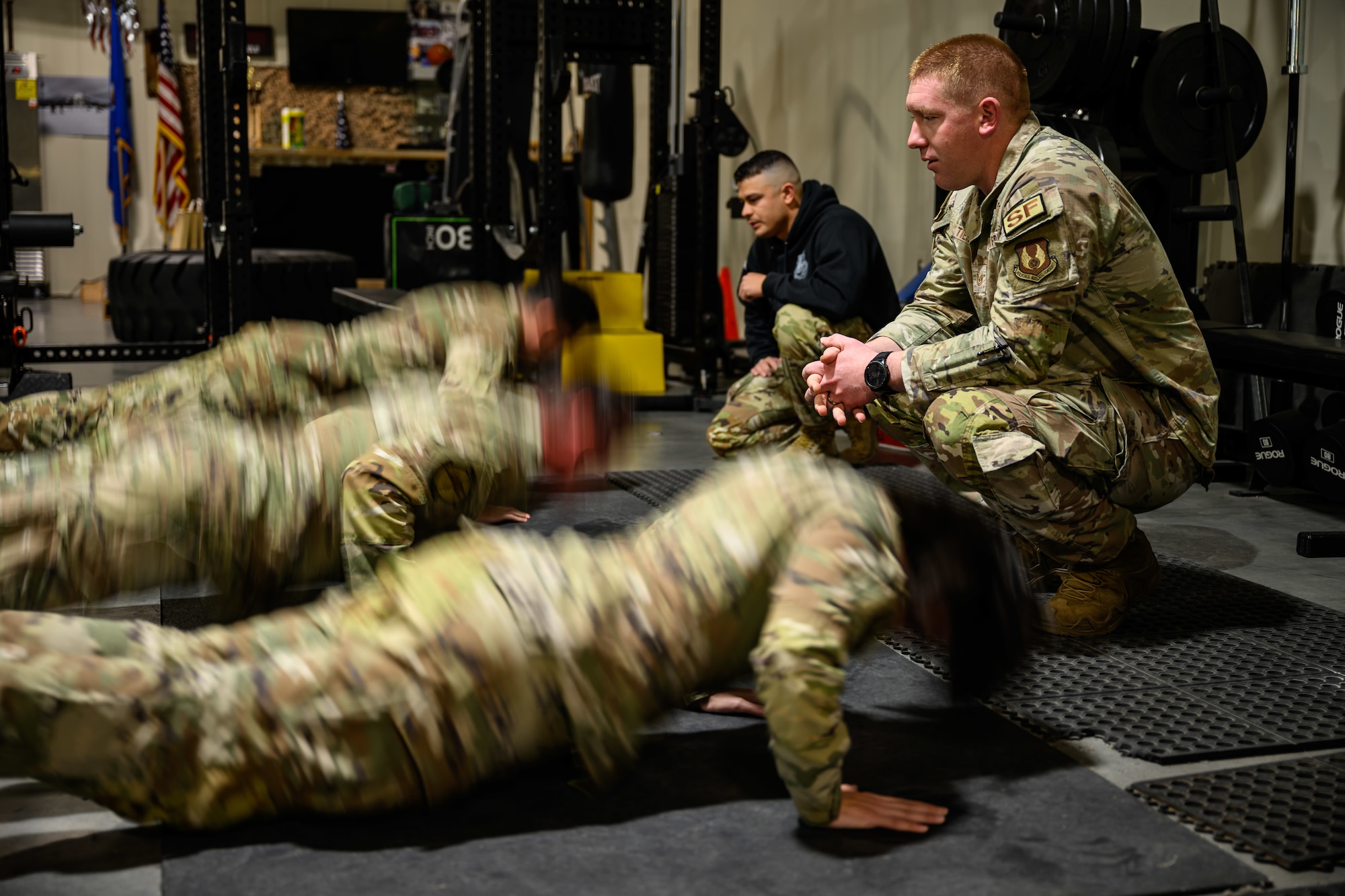 Tech. Sgt. James Tienor crouches down at right watching Airmen doing pushups at left.