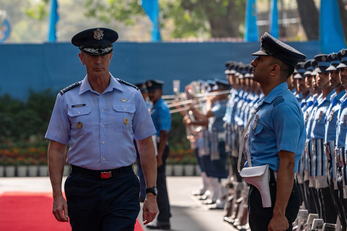 U.S. Air Force Gen. Kevin Schneider walks on a red carpet, inspecting a line of uniformed service members standing at attention. A military band is visible in the background.