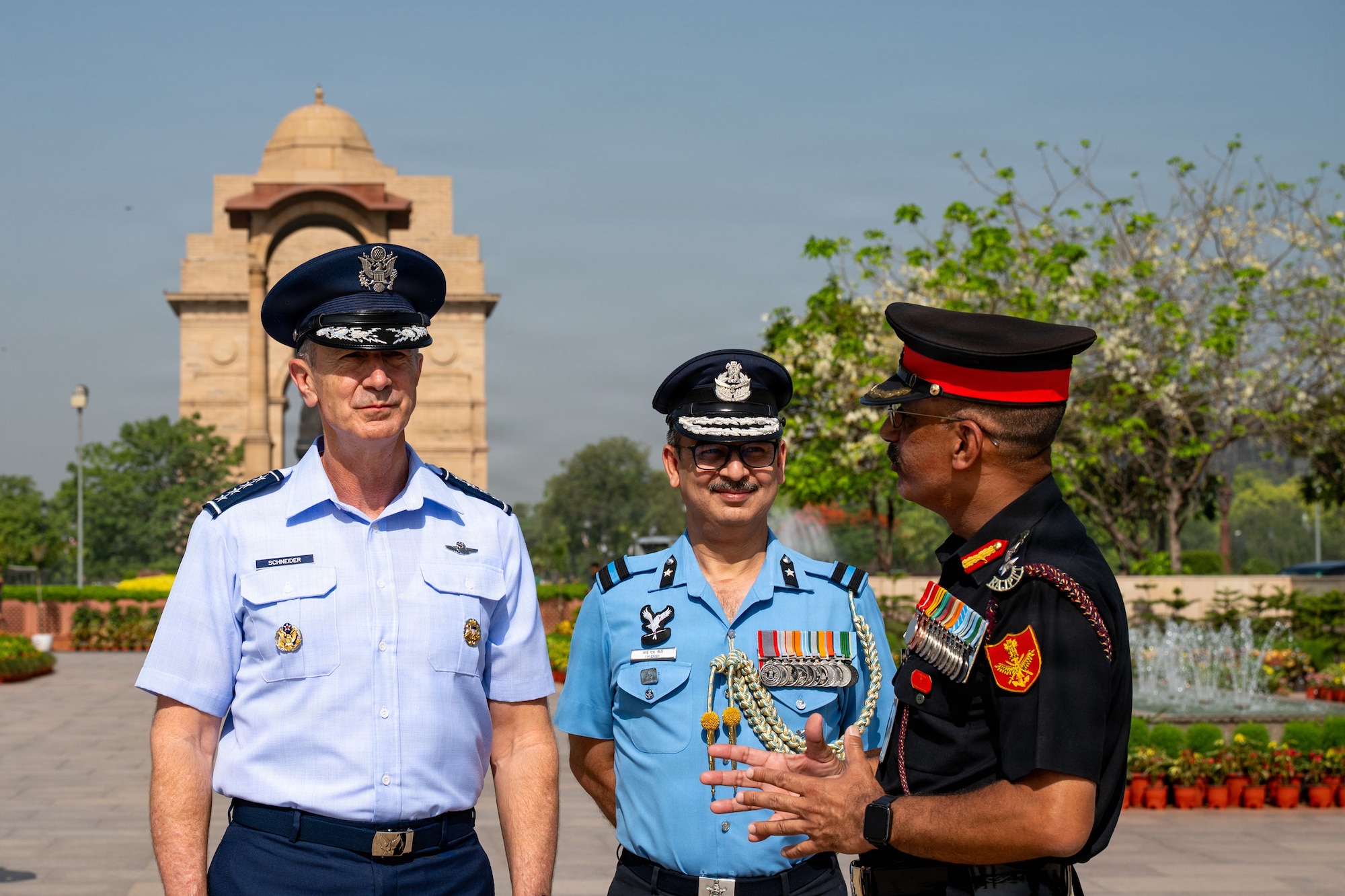 U.S. Air Force Gen. Kevin Schneider and two members of the Indian Armed Forces, each in different service uniforms, stand together outdoors. One Indian officer gestures while speaking, with a large stone archway visible in the background.