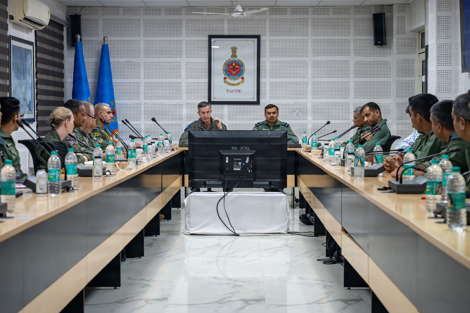 USAF Gen. Kevin Schneider, PACAF commander, and another leader sit at the head of a conference table briefing a group of military pilots in flight suits. Behind them are flags and a TACDE wall emblem.
